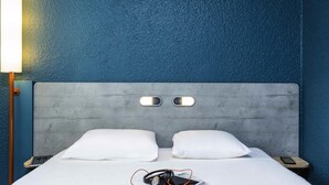 A hotel room with a grey headboard, two white pillows, a bedside table with a lamp, a smartphone, and headphones. - ibis budget Paris Porte d'Italie Est (Le Kremlin-Bicetre)