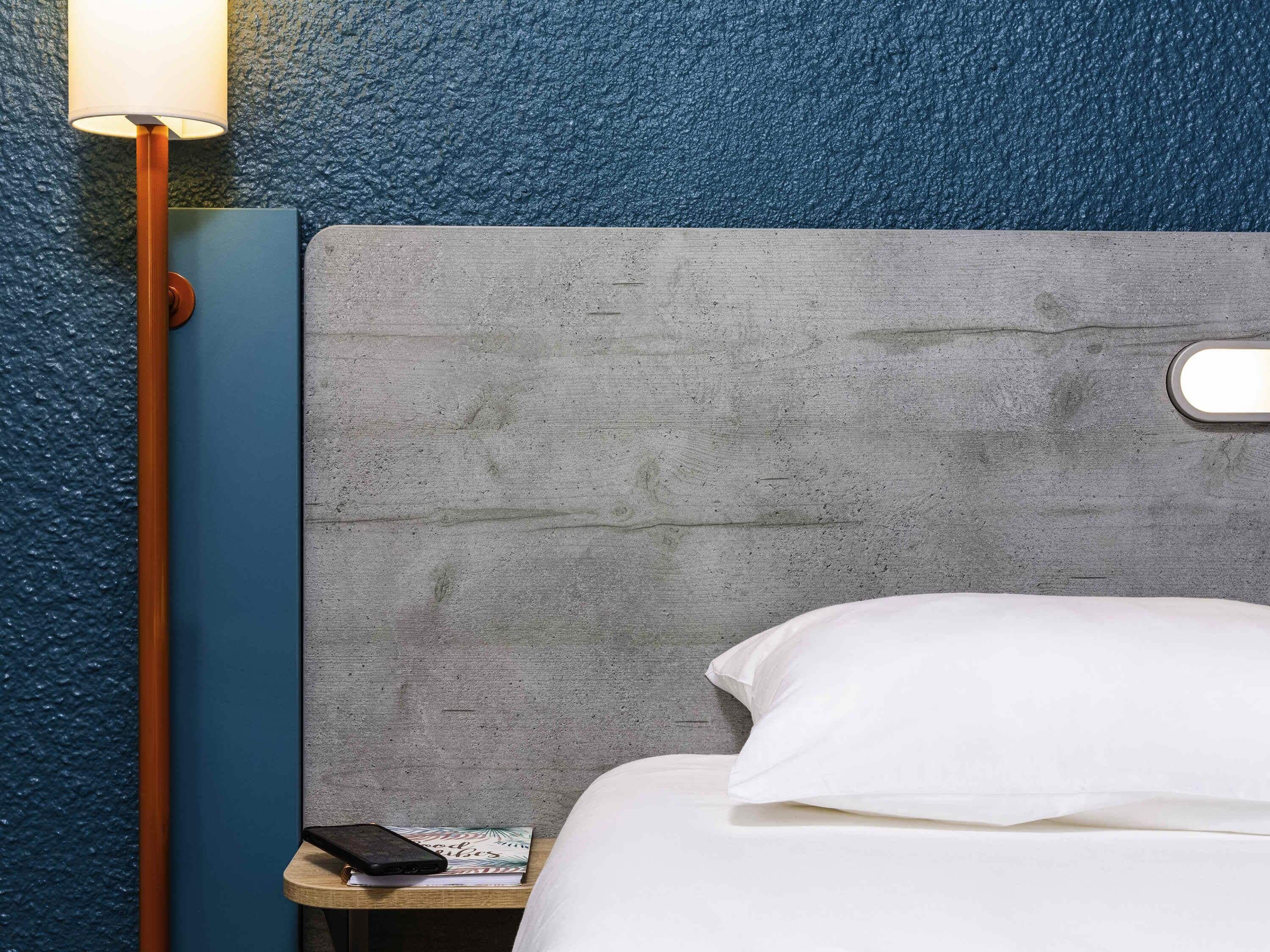 A bed with white bedding, a grey headboard, a bedside table with a lamp, and a book.