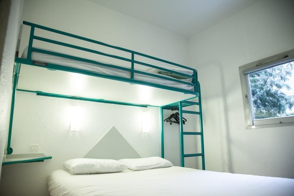 A bunk bed room with a white mattress, teal railings, a triangular wall decoration, and a window with a view of trees.
