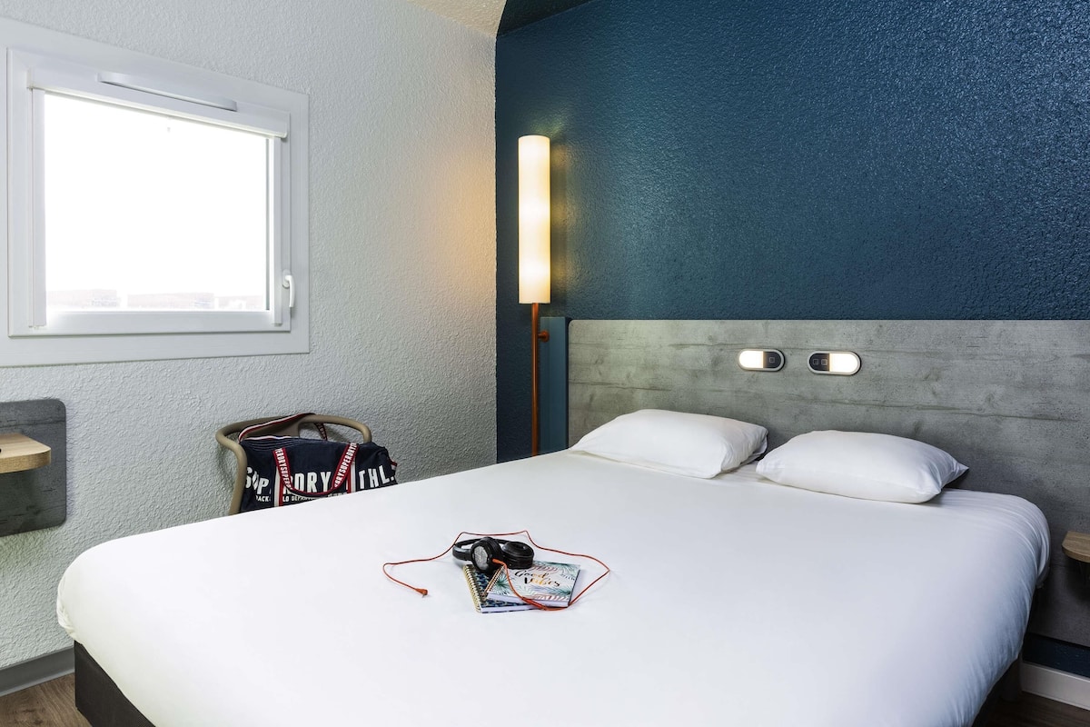 A hotel room with a bed, a bedside table, a lamp, a window, and a bag on the floor.