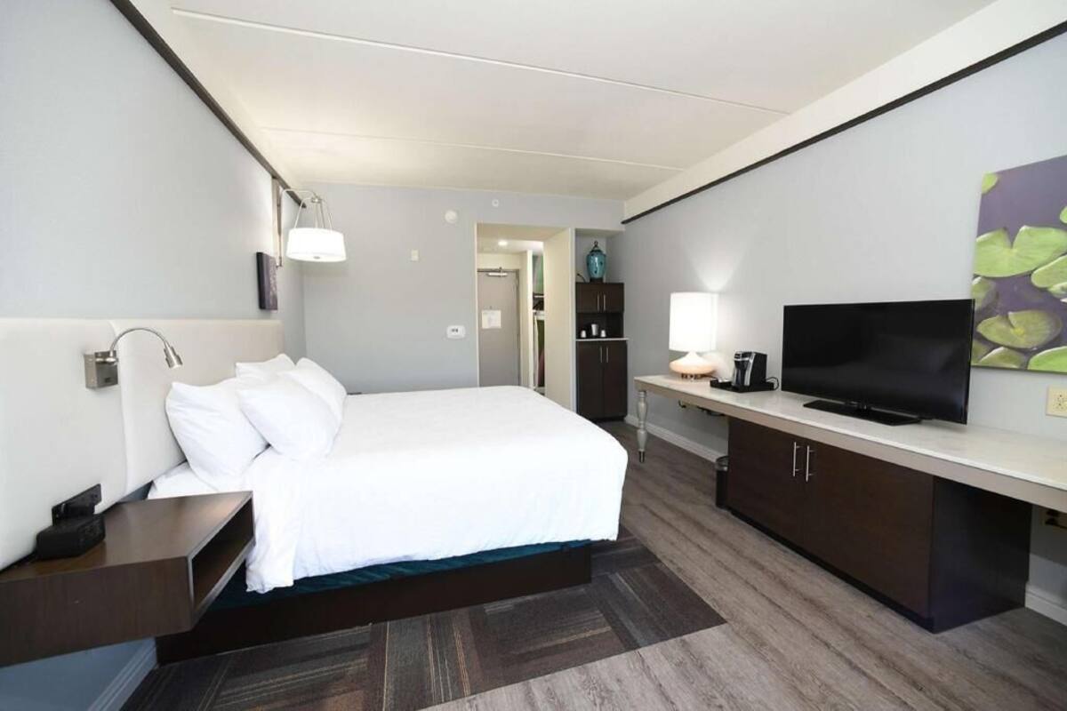 A hotel room with a bed, bedside table, lamp, TV, and a cabinet.