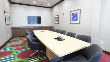 A conference room with a long table, six chairs, a flat-screen TV on the wall, and colorful carpet.