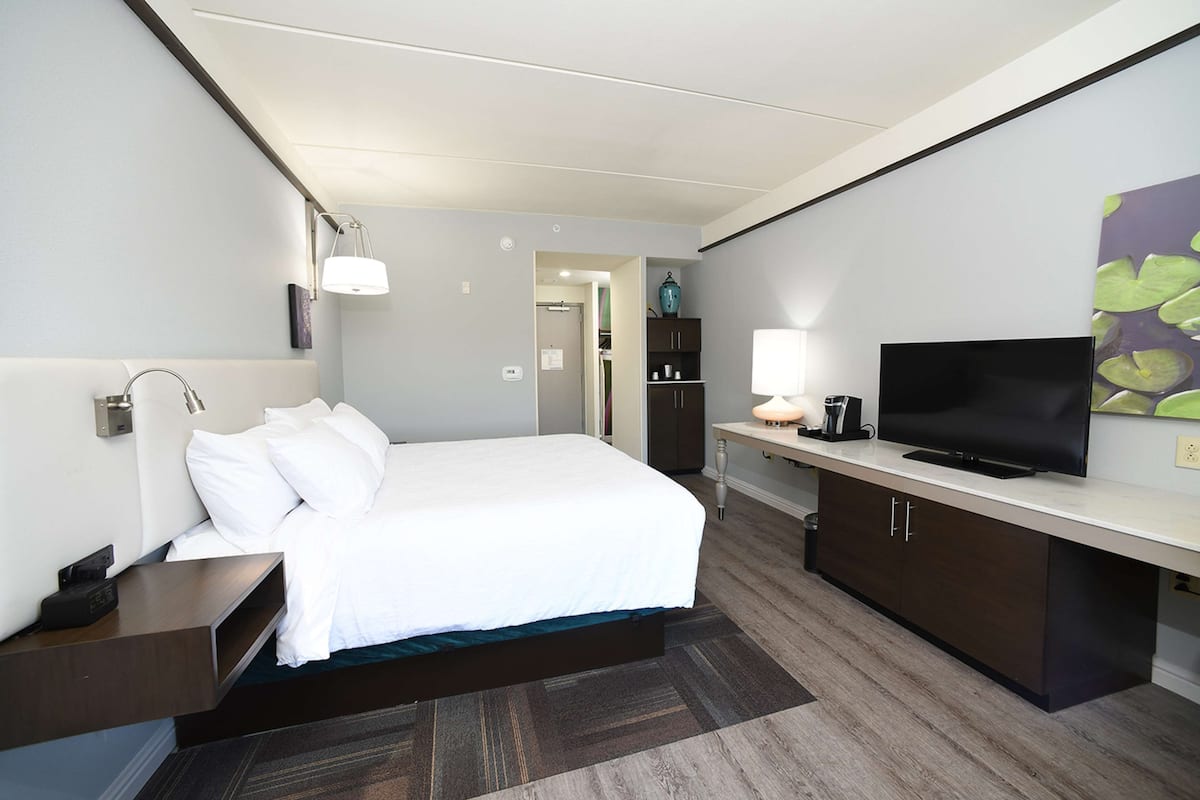 A hotel room with a bed, bedside table, lamp, TV, wardrobe, and a painting on the wall.