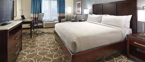 Quarto de hotel com cama grande, escrivaninha com cadeira, televisão e janela com cortinas.