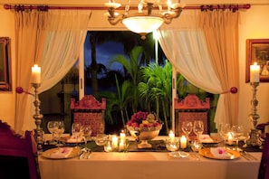 A dining table set with candles, glasses, and a fruit bowl, with curtains and a chandelier in the background.