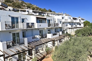 Una hilera de edificios blancos con balcones, algunos con pérgolas de madera, y una colina al fondo.