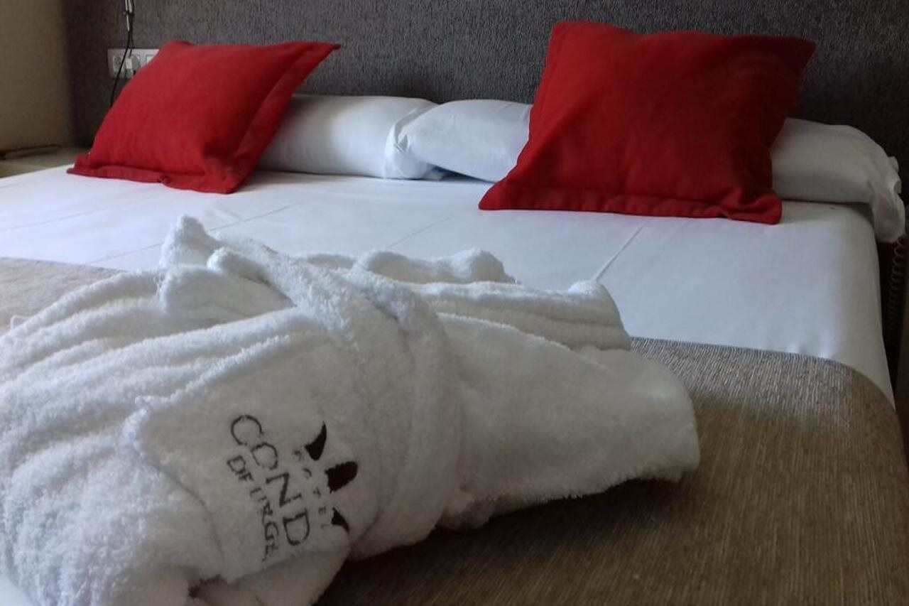 Una cama bien hecha con sábanas blancas y almohadas rojas, una bata blanca con texto estampado, y un cabecero de madera.