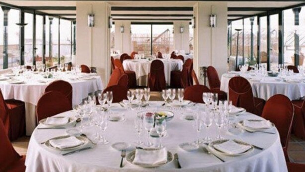 A restaurant with round tables set for dining, red chairs, and a view through large windows. - Condes de Urgel (Lleida)