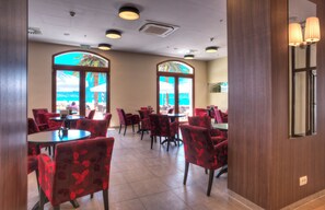 A hotel dining area with red upholstered chairs and tables, large windows with a view of the beach, and a wooden wall with a mirror. - Pine (Tivat)