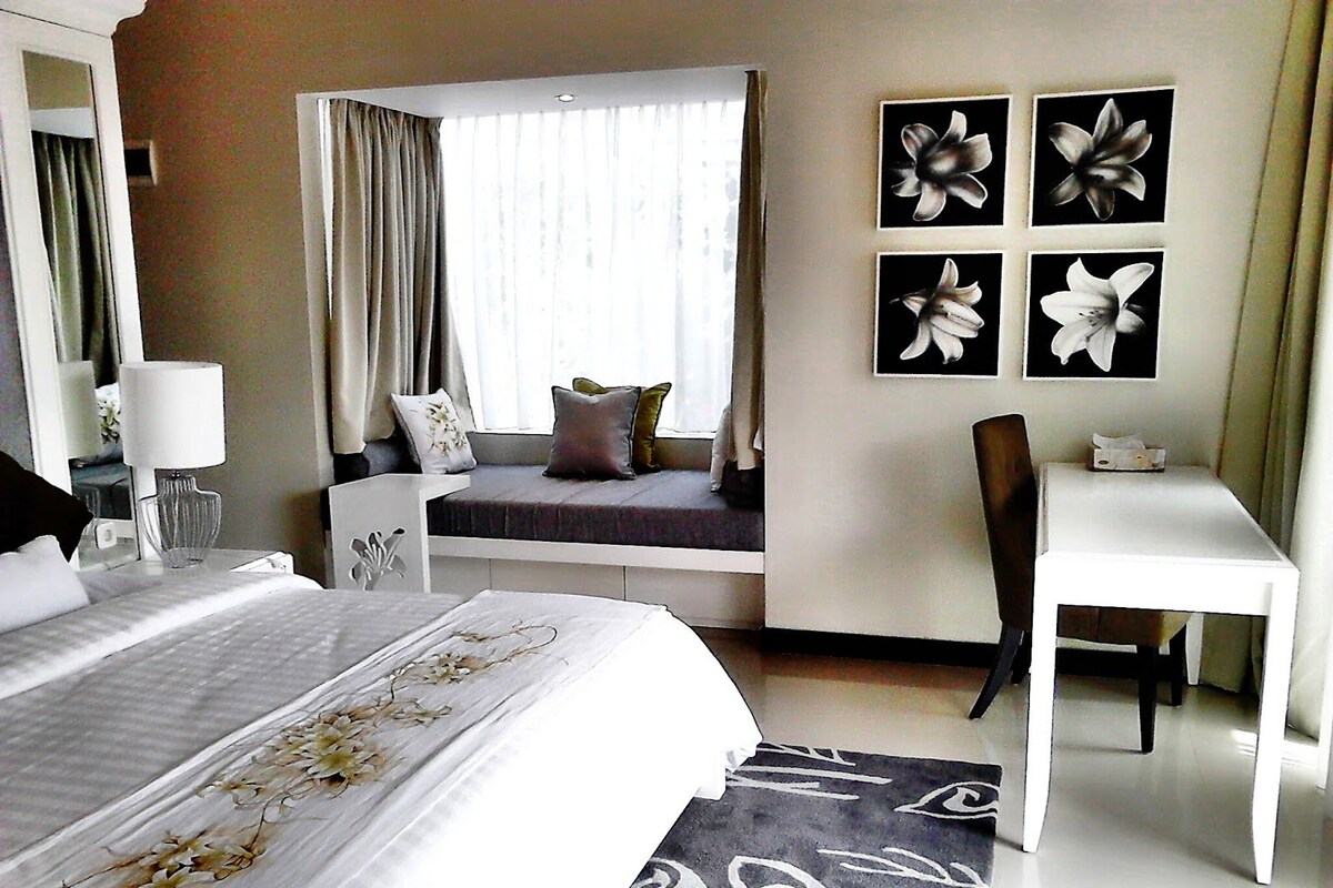 A hotel room with a bed, a desk, a chair, a window with curtains, and wall art.
