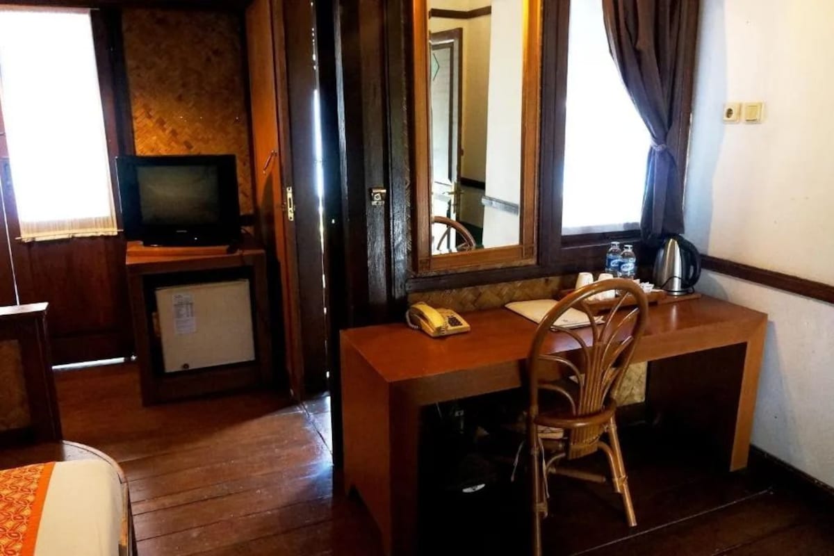 A hotel room with a wooden desk, a chair, a television, a small refrigerator, and a mirror.
