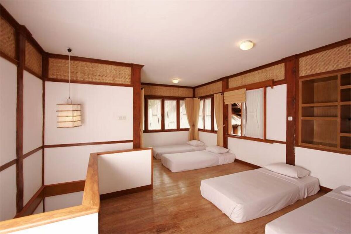 Una habitació tradicional japonesa amb sòls de tatami, bigues de fusta i panells shoji. Hi ha tres esteves de tatami amb roba de llit blanca, una làmpada penjant i una finestra amb vistes.