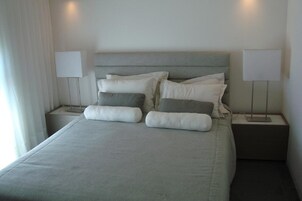 Una cama bien hecha con cabecera gris, dos lámparas de noche y una ventana con cortinas.