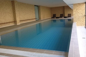 An indoor swimming pool with clear blue water, surrounded by beige textured walls and a tiled floor.