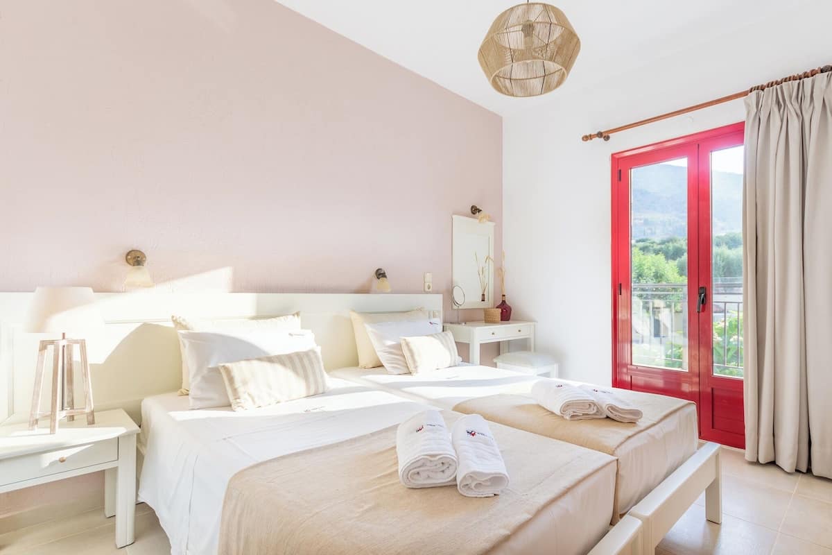 A hotel room with a bed, bedside tables, a mirror, and a view of greenery through a red-framed window.