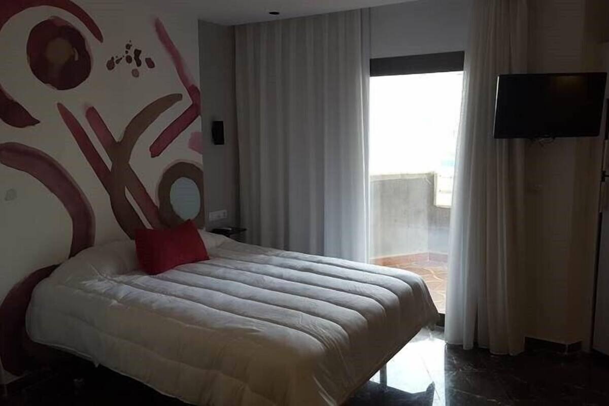 A modern hotel room with a large bed, a flat-screen TV, and a balcony with curtains.