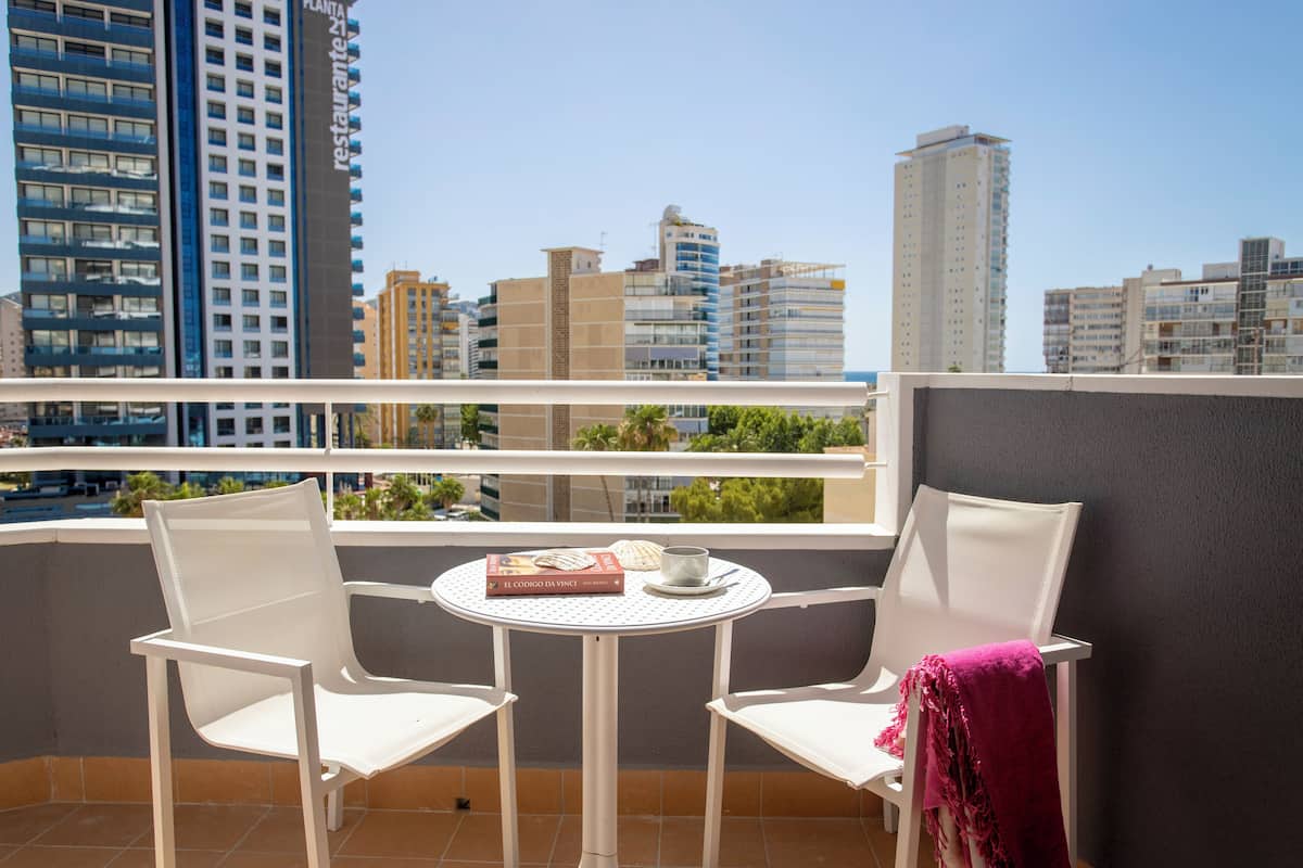 A balcony with two white chairs, a small round table with a book and a cup, and a view of high-rise buildings.