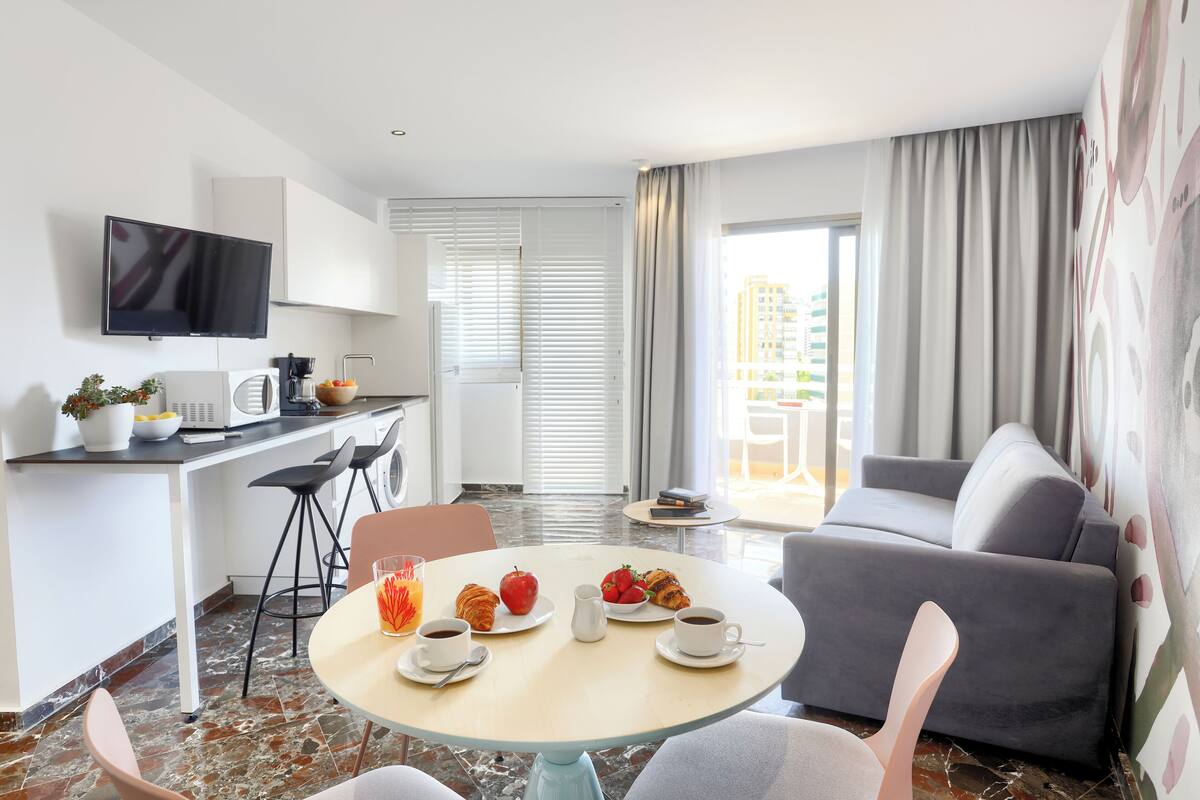 A modern hotel room with a dining area, a sofa, a TV, and a balcony with curtains.