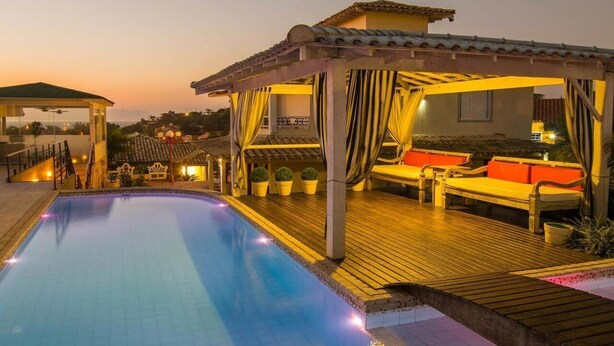 A poolside area with a wooden deck, a covered seating area with cushions, and a building with a tiled roof in the background. - Pousada Aroma do Mar (Búzios)