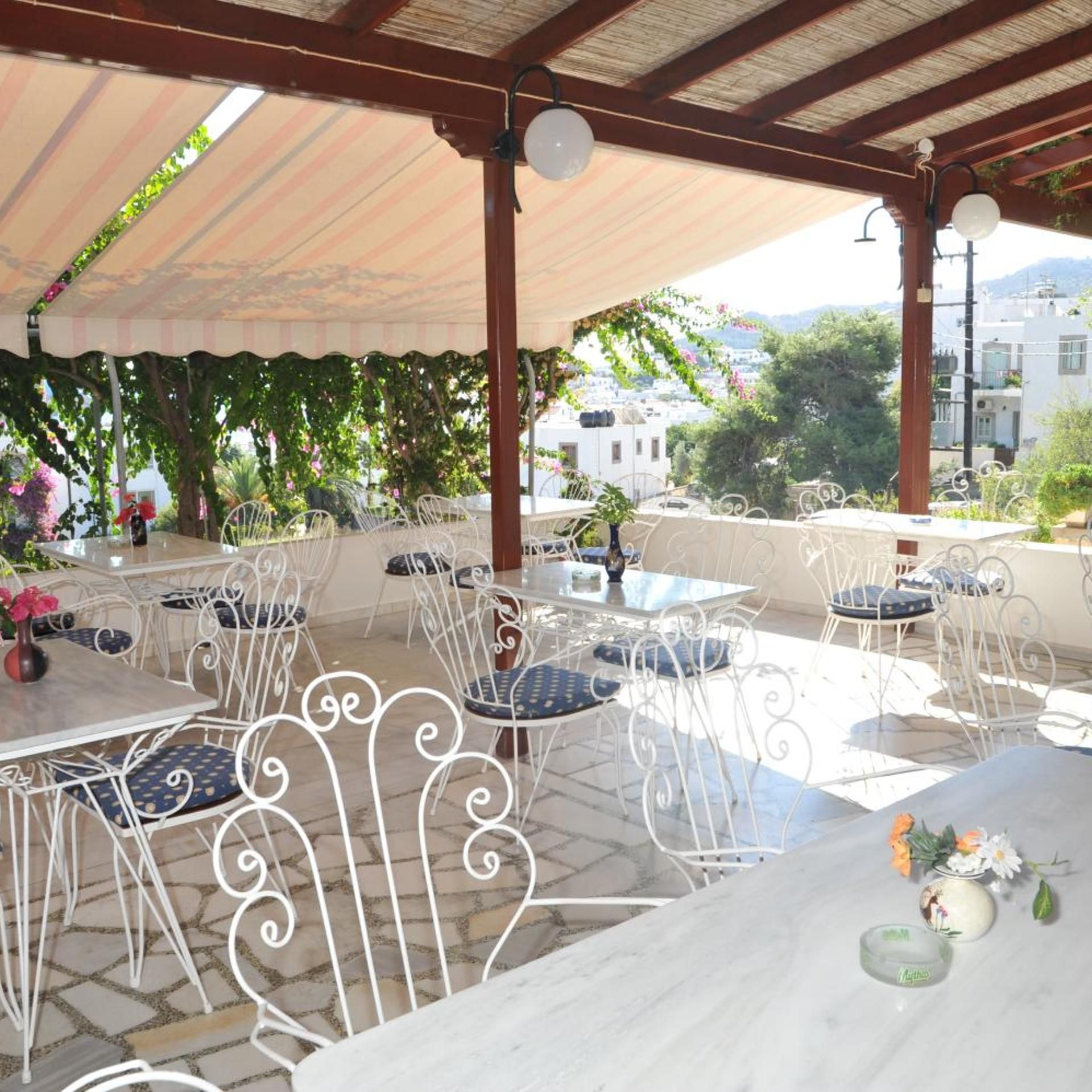 An outdoor seating area with white metal chairs and tables, covered by a striped awning, and overlooking a residential area with trees and buildings.