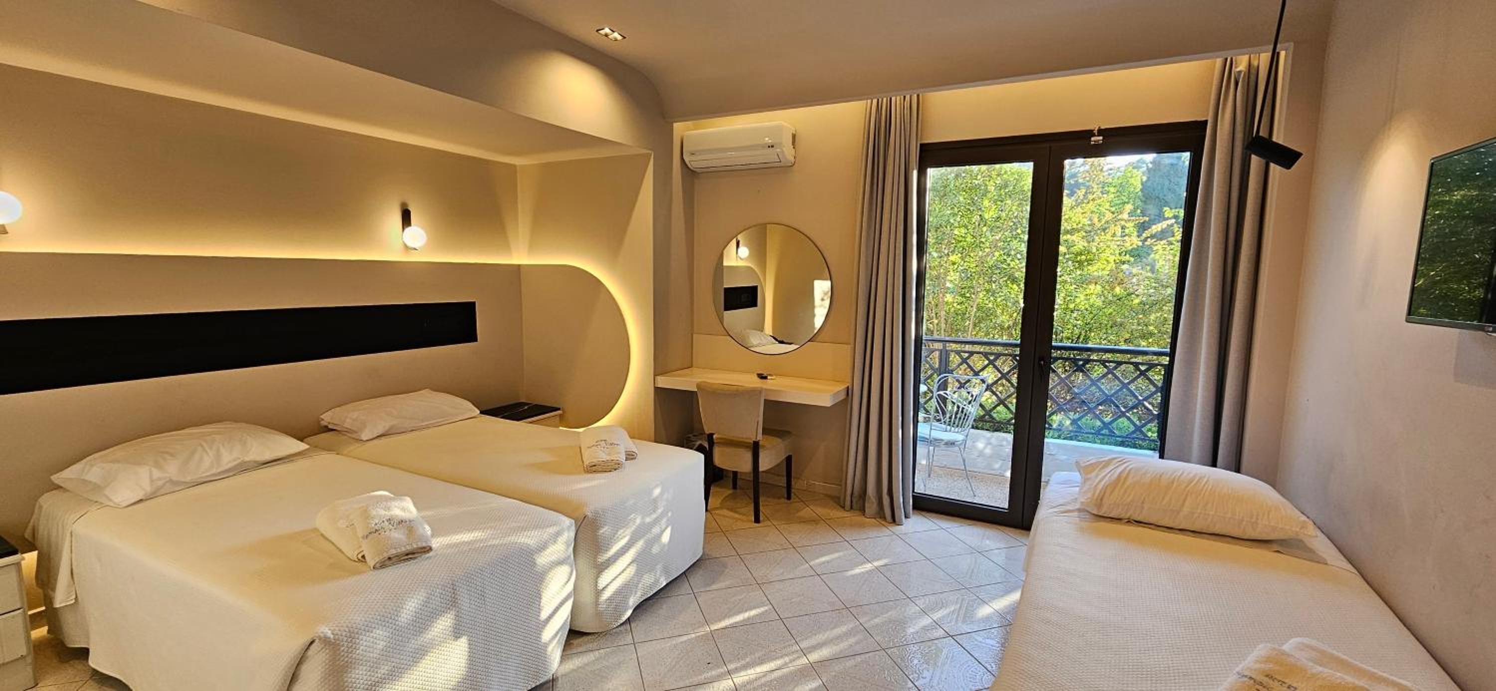 Camera d'albergo con due letti, una scrivania, uno specchio e un balcone con vista sul verde.