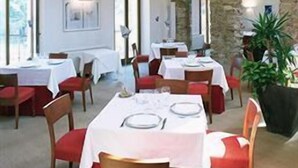 Un comedor con mesas dispuestas para dos, vigas de madera en el techo, paredes de piedra y ventanas amplias.