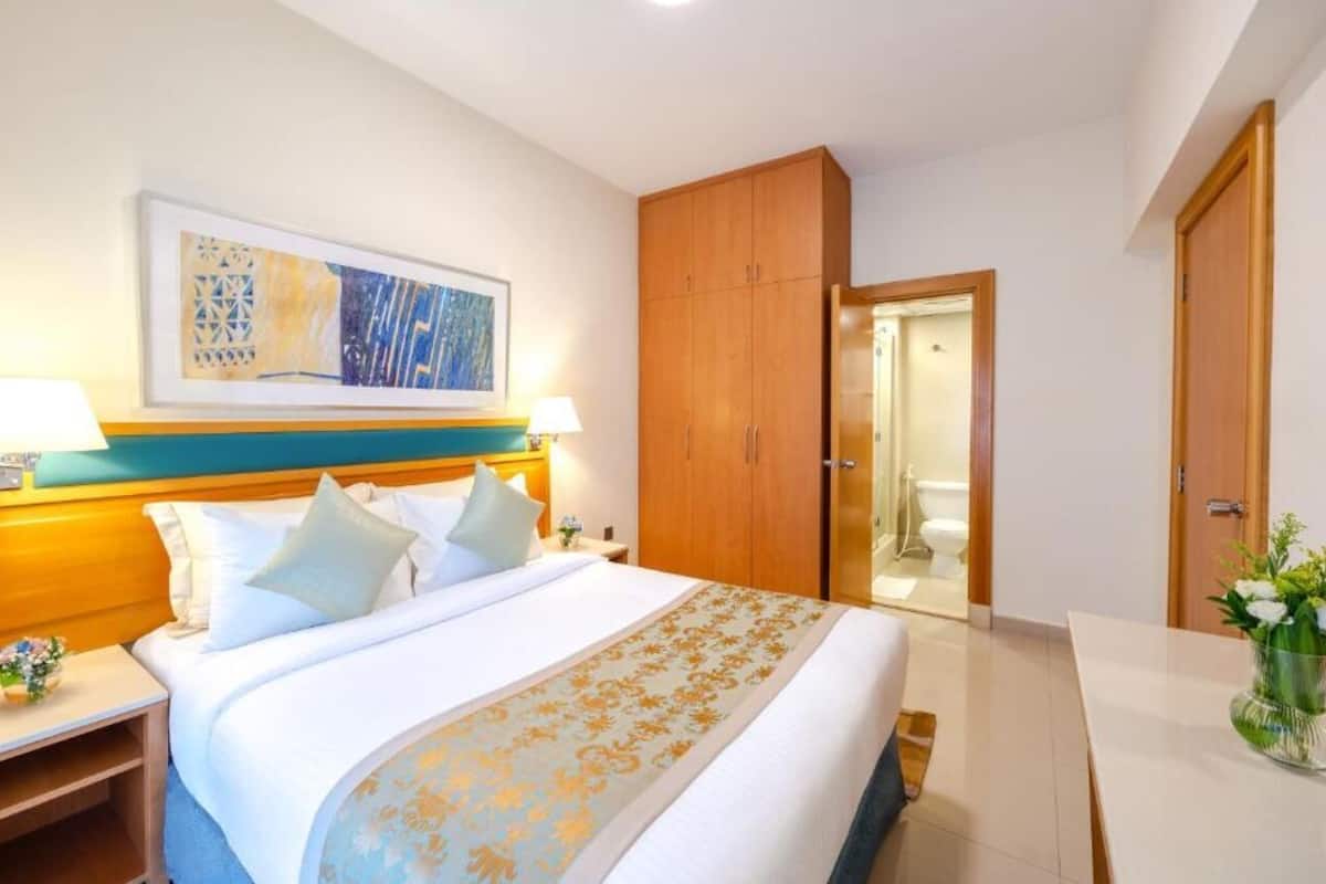 A hotel room with a large bed, wooden wardrobe, bedside tables with lamps, and a framed artwork on the wall.