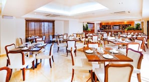 Un comedor amplio con varias mesas y sillas, una zona de bar con televisión y ventanas grandes con persianas.