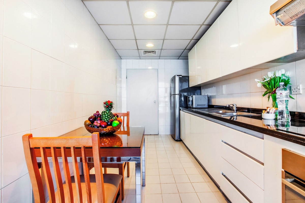 A compact kitchen with white cabinets, a stainless steel refrigerator, and a wooden dining table with a fruit bowl.