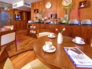 Habitación de hotel con piso de madera, una mesa de comedor con sillas blancas, una estación de café y un reloj en la pared.