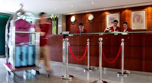 Un mostrador de recepción de hotel con una mampara de vidrio, un reloj y retratos enmarcados en la pared.