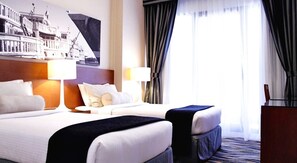 A hotel room with two beds, a headboard, a nightstand, a lamp, a window with curtains, and a wall-mounted artwork.