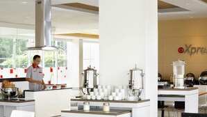 A modern kitchen with a chef preparing food, coffee machines, and a branded wall. - Amaris Hotel Tebet (Jakarta)