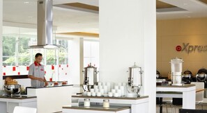 A modern kitchen with a chef preparing food, coffee machines, and a branded wall. - Amaris Hotel Tebet (Jakarta)