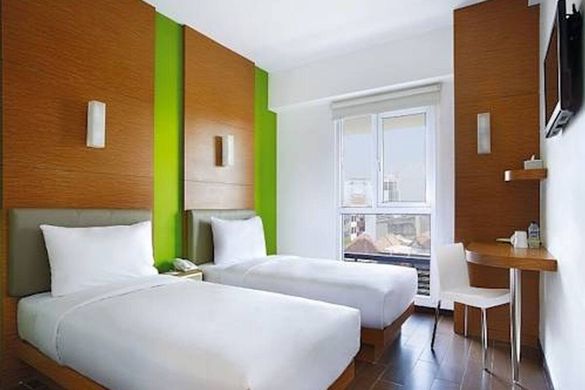 A hotel room with two beds, a desk, a chair, a TV, and a window.