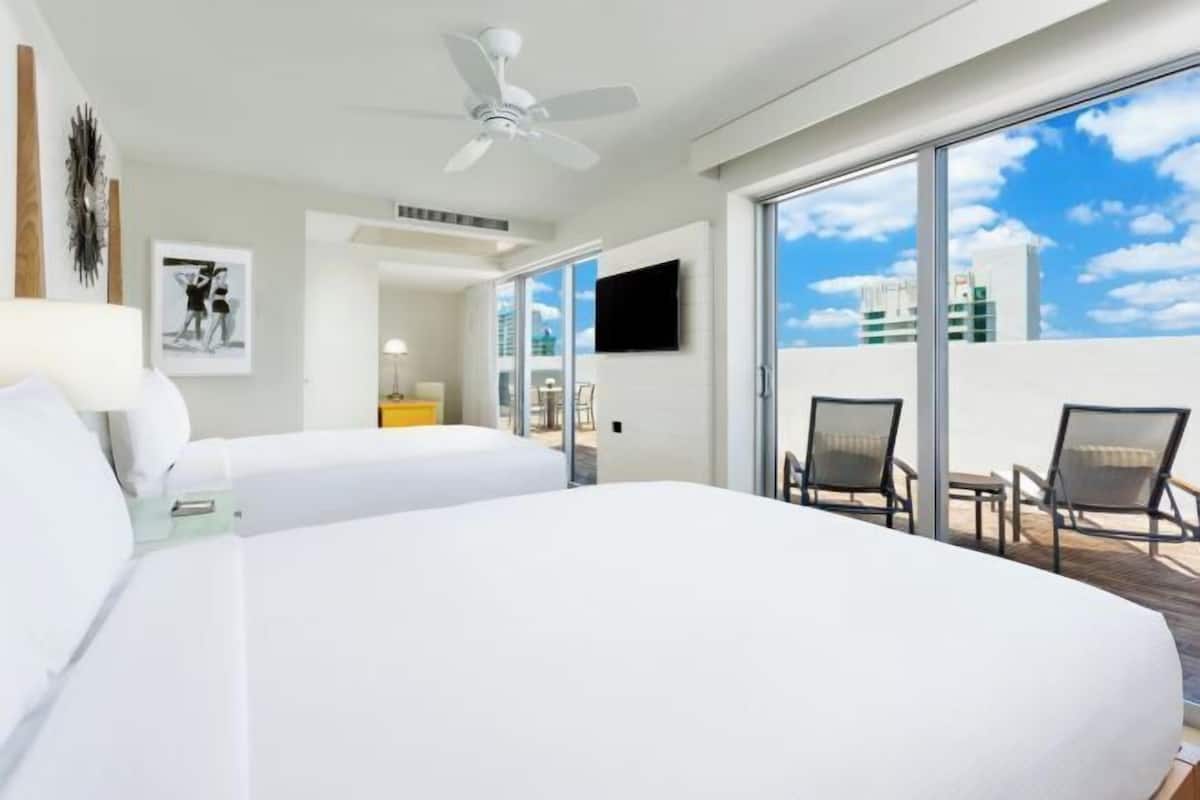 Habitación de hotel con dos camas, ventilador de techo, televisor y balcón con sillas.