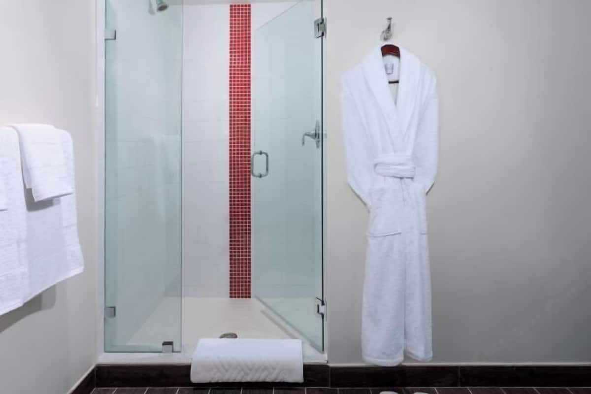 Une douche en verre avec des carreaux rouges en guise de décoration, un peignoir blanc suspendu à un porte-serviettes et une serviette blanche sur le sol.