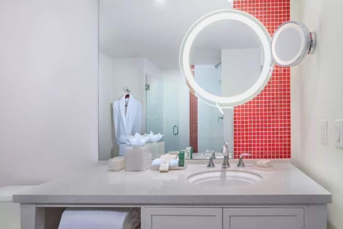 Un baño moderno con lavabo doble, un espejo grande con luz circular y pared de azulejos rojos.