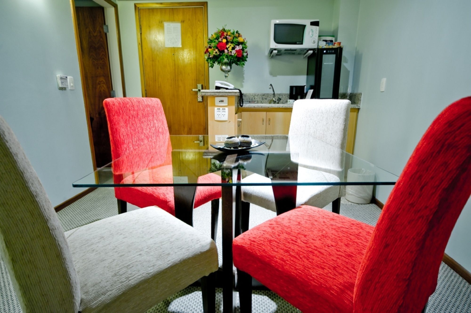 Uma sala de jantar com mesa de vidro e quatro cadeiras, duas vermelhas e duas brancas, num espaço com porta de madeira e micro-ondas ao fundo.