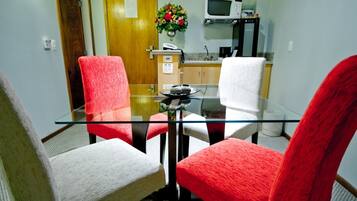Uma sala de jantar com mesa de vidro e quatro cadeiras, duas vermelhas e duas brancas, num espaço com porta de madeira e micro-ondas ao fundo.