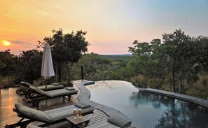 Zona junto a la piscina con sillones de descanso y una mesa, rodeada de exuberante vegetación y vistas al paisaje.