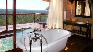 A bathroom with a large bathtub, a sink, and a mirror. There is a view of a lush landscape through large windows.