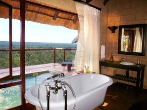 Un baño con una bañera grande, un lavamanos y un espejo. Se ve un paisaje exuberante a través de amplias ventanas.