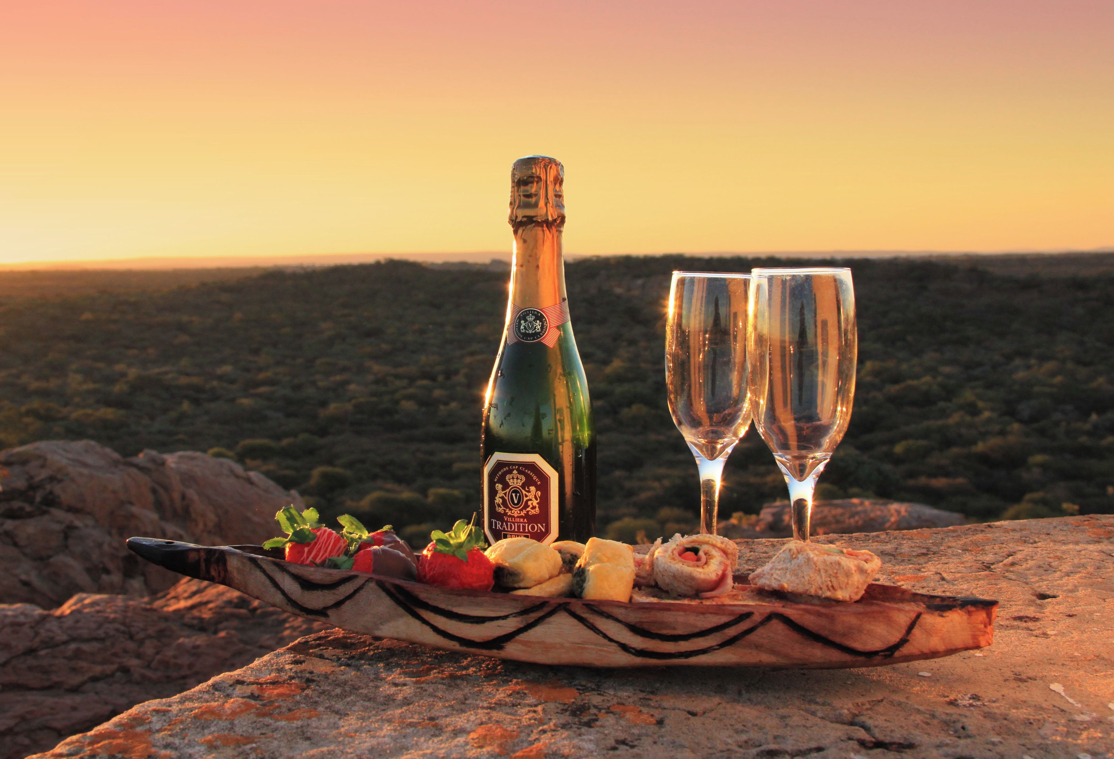 A bottle of wine and two glasses on a wooden board with snacks, set against a scenic sunset background.