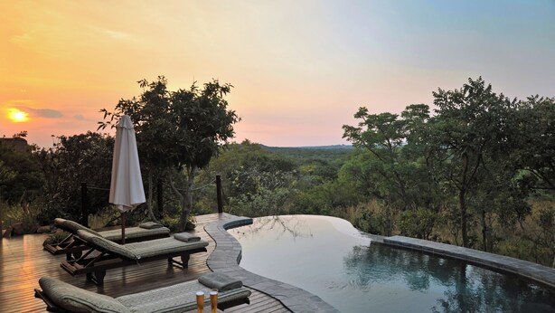 Uma área à beira da piscina com espreguiçadeiras e uma mesa, rodeada por vegetação exuberante e vista para a paisagem.