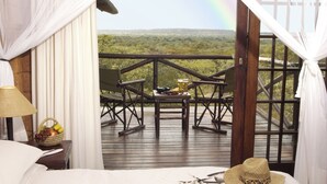 Quarto com cama, mesa de cabeceira com luminária e cesta de frutas, deck de madeira com cadeiras e mesa, e vista para uma paisagem com arco-íris.