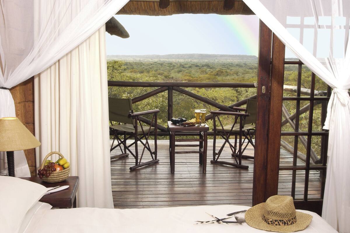 Quarto com cama, mesa de cabeceira com luminária e cesta de frutas, deck de madeira com cadeiras e mesa, e vista para uma paisagem com arco-íris.