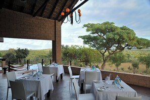 Un área de comedor al aire libre con mesas preparadas para los huéspedes, con vistas a un paisaje escénico con árboles y colinas.