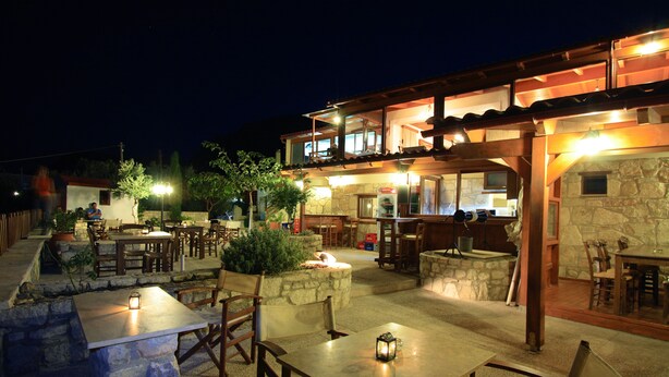 An outdoor patio area with stone tables and wooden chairs, a stone fireplace, and a wooden pergola structure. - Earino (Heraklion)