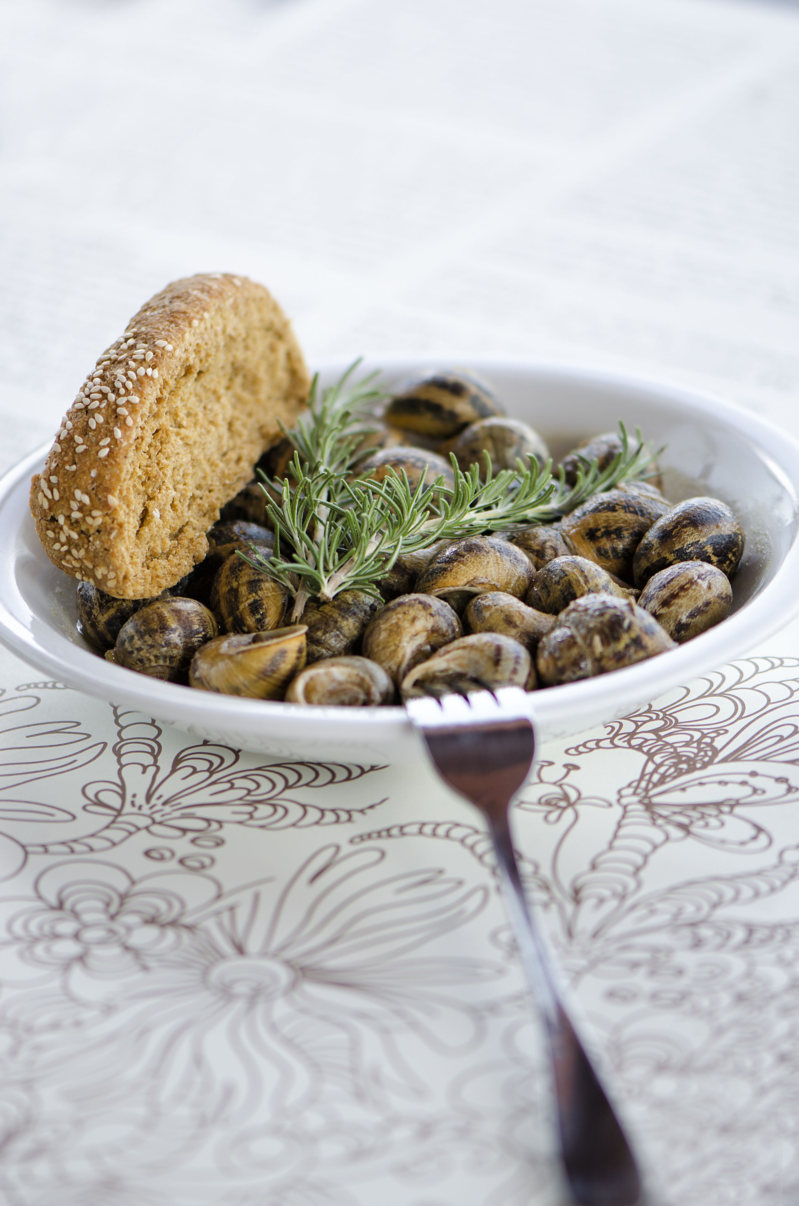 A bowl of mushrooms with a slice of bread and rosemary on a patterned tablecloth.